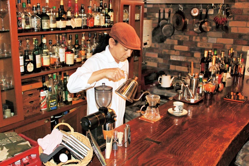 Mikaduki's owner pours a v60 filter coffee behind the bar in one of signature flat caps, a wall of Japanese and global whiskey is behind him