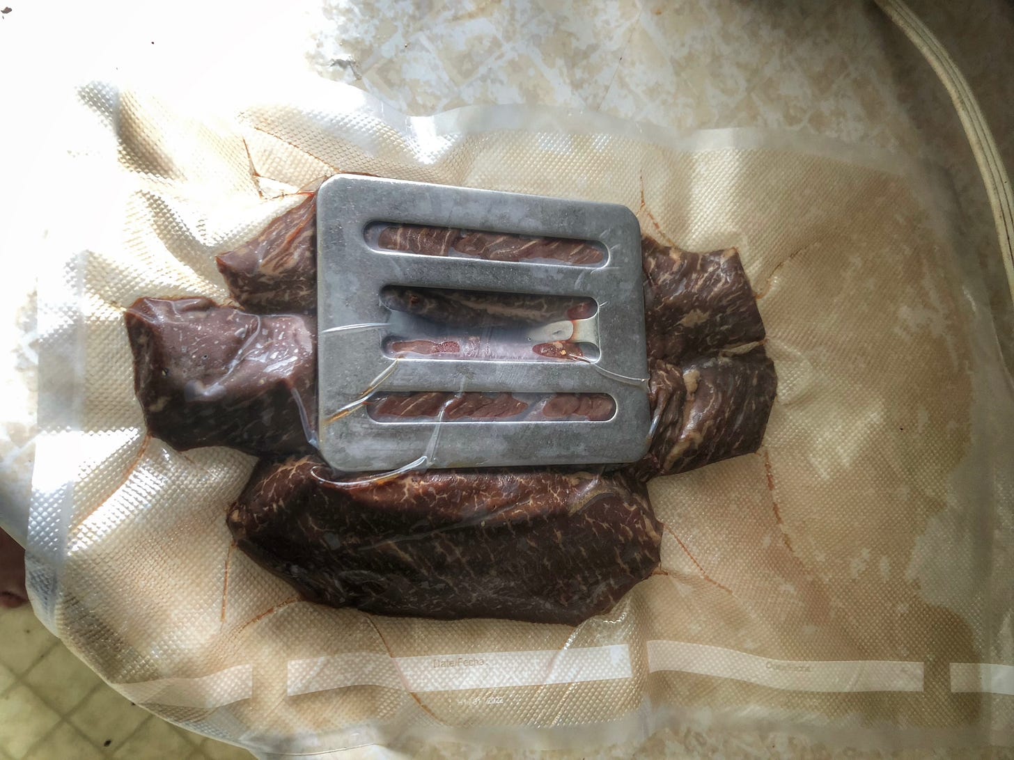 A vacuum sealed bag of boneless short ribs, ready to go into a sous vide bath to cook. A Chef's Press is sealed inside the bag with the meat to help keep it submerged.
