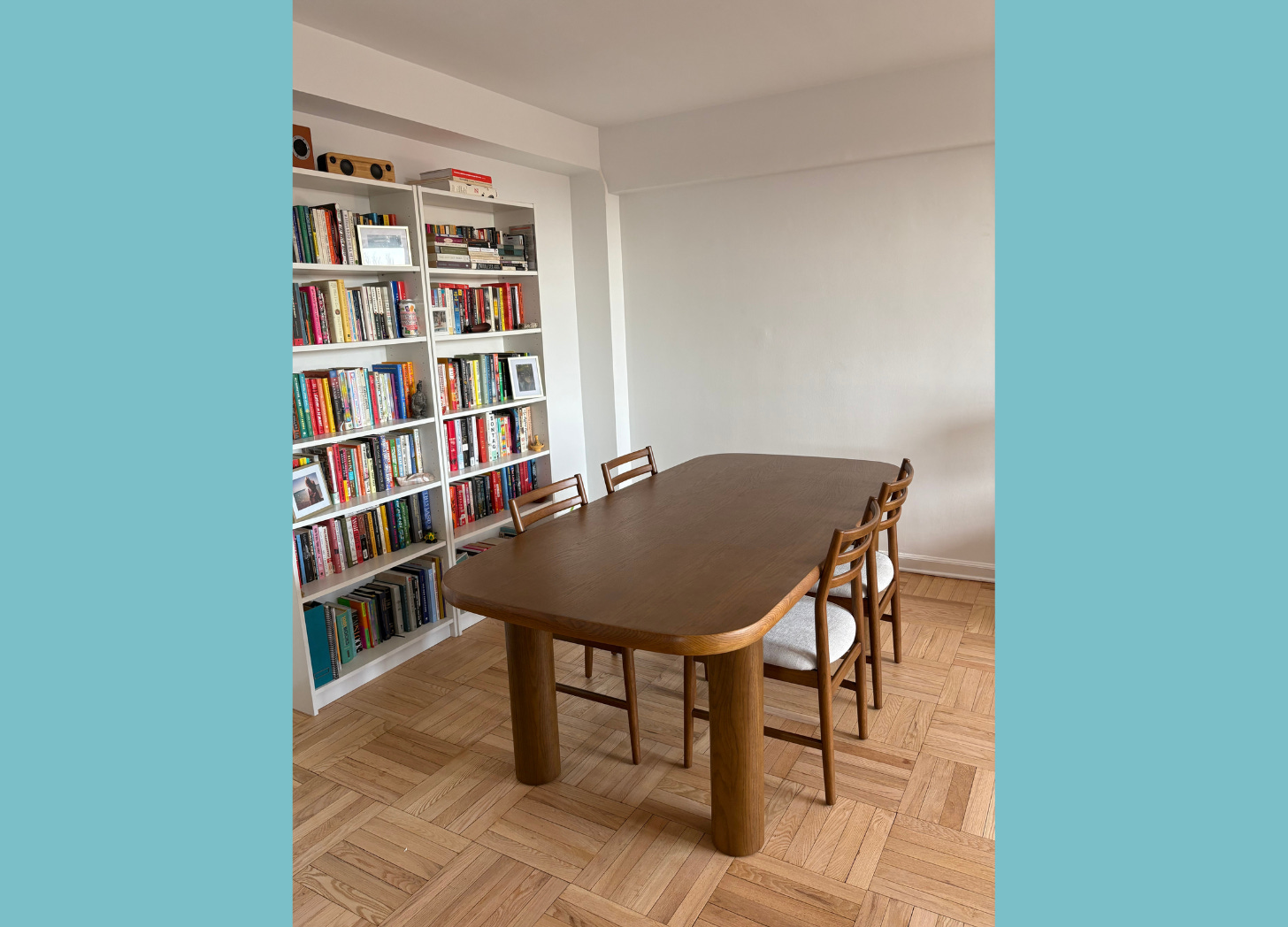Photo of a dining table with four chairs and a bookcase filled with books in the background.