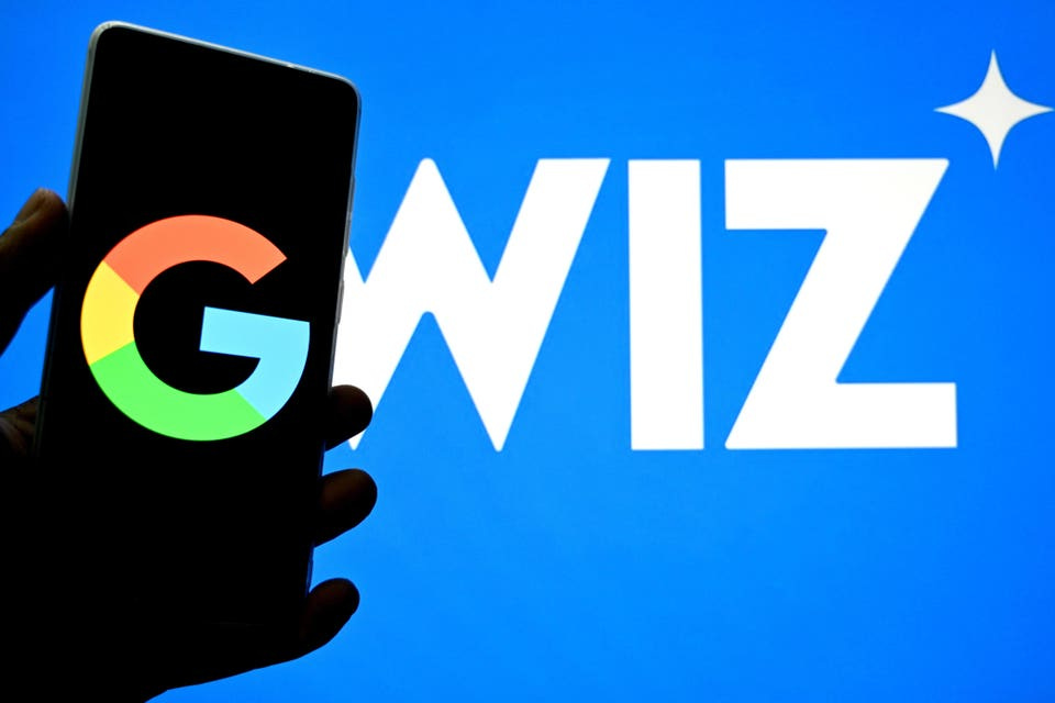 Smartphone displaying Google’s logo held up against background featuring the Wiz logo Smartphone displaying Google’s logo held up against background featuring the Wiz logo