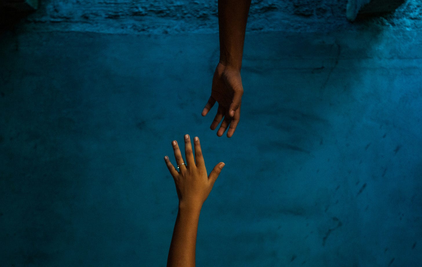 Two hands reaching toward each other against a textured blue background, one extended from above and the other from below, symbolising connection and support. Two hands reaching toward each other against a textured blue background, one extended from above and the other from below, symbolising connection and support.