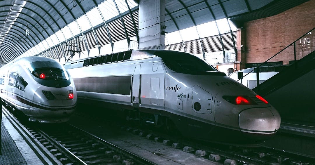 a silver train traveling down train tracks next to a loading platform a silver train traveling down train tracks next to a loading platform