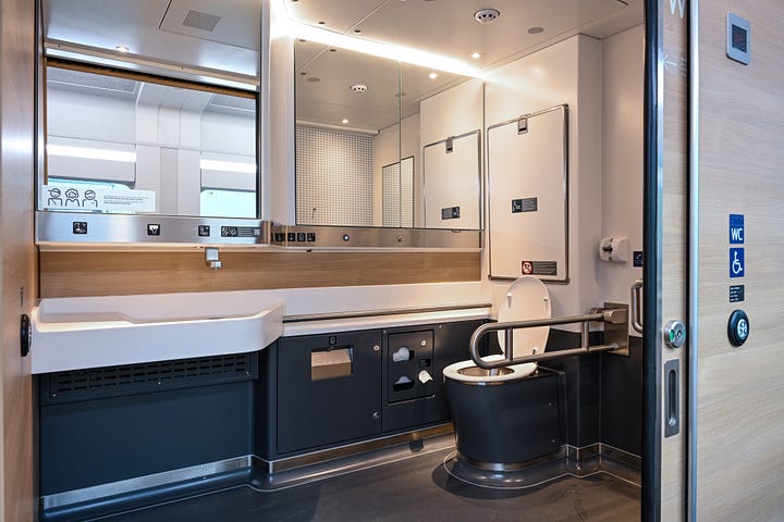 1. Image: White train from above, 2. Interior of the train, blue seats and wooden tables, 3. wheelchair space with an empty wheelchair and a wooden table 4. Accessible toilet with a lot of mirrors, a wide door and grabrails 