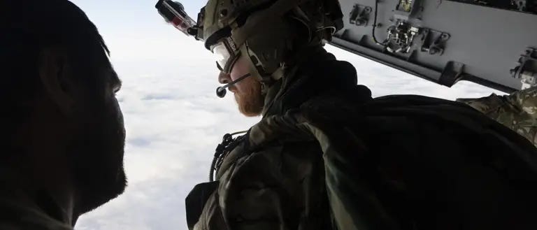 Two individuals in military gear are inside an aircraft, looking out over a cloudy sky. One person is wearing a helmet with a camera, gear, and patches with the UK flag. The interior of the aircraft shows various equipment and panels.
