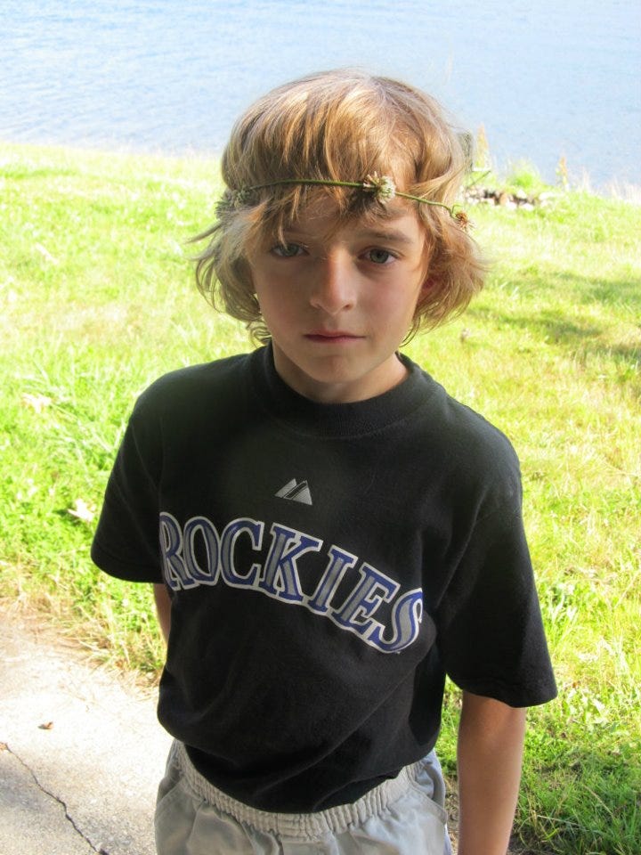 A small blonde boy at the water's edge, wearing a flower crown, a Rockies t-shirt, and a melancholy expression