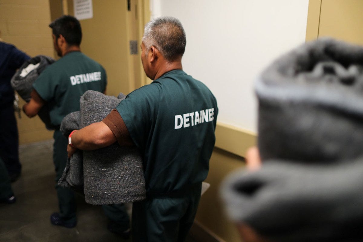Detainees, wearing green jumpsuits and holding gray blankets stand in line, inside a detention facility. Detainees, wearing green jumpsuits and holding gray blankets stand in line, inside a detention facility.
