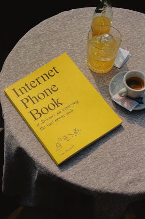 L'Internet Phone Book posé sur une table de bistrot, à côté d'un café. Sa couleur et sa typographie rappelle en effet les vieux annuaires téléphoniques des pages jaunes