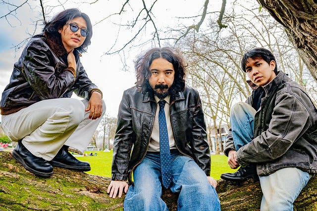 Three Asian men in jeans and dark jackets sitting on a large tree branch in a grassy field