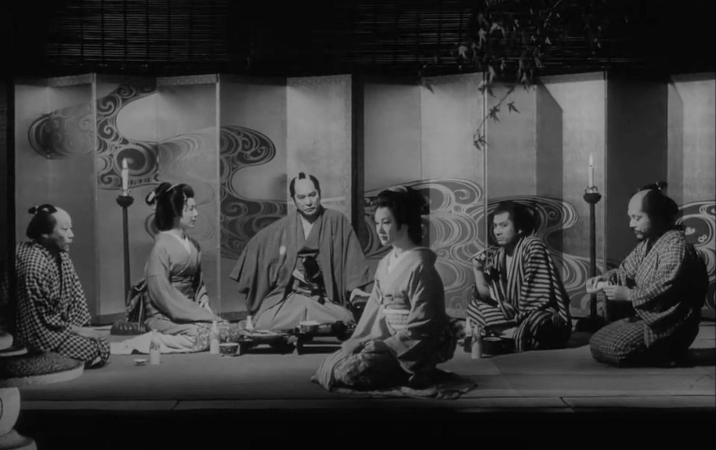 Film still from the 1971 movie “Demons/Shura”. 4 Japanese men and 2 Japanese women in kimonos and hakatas sit in front of a folding screen with a wave design. Everyone is staring at one of the women who is seated closer to the viewer.