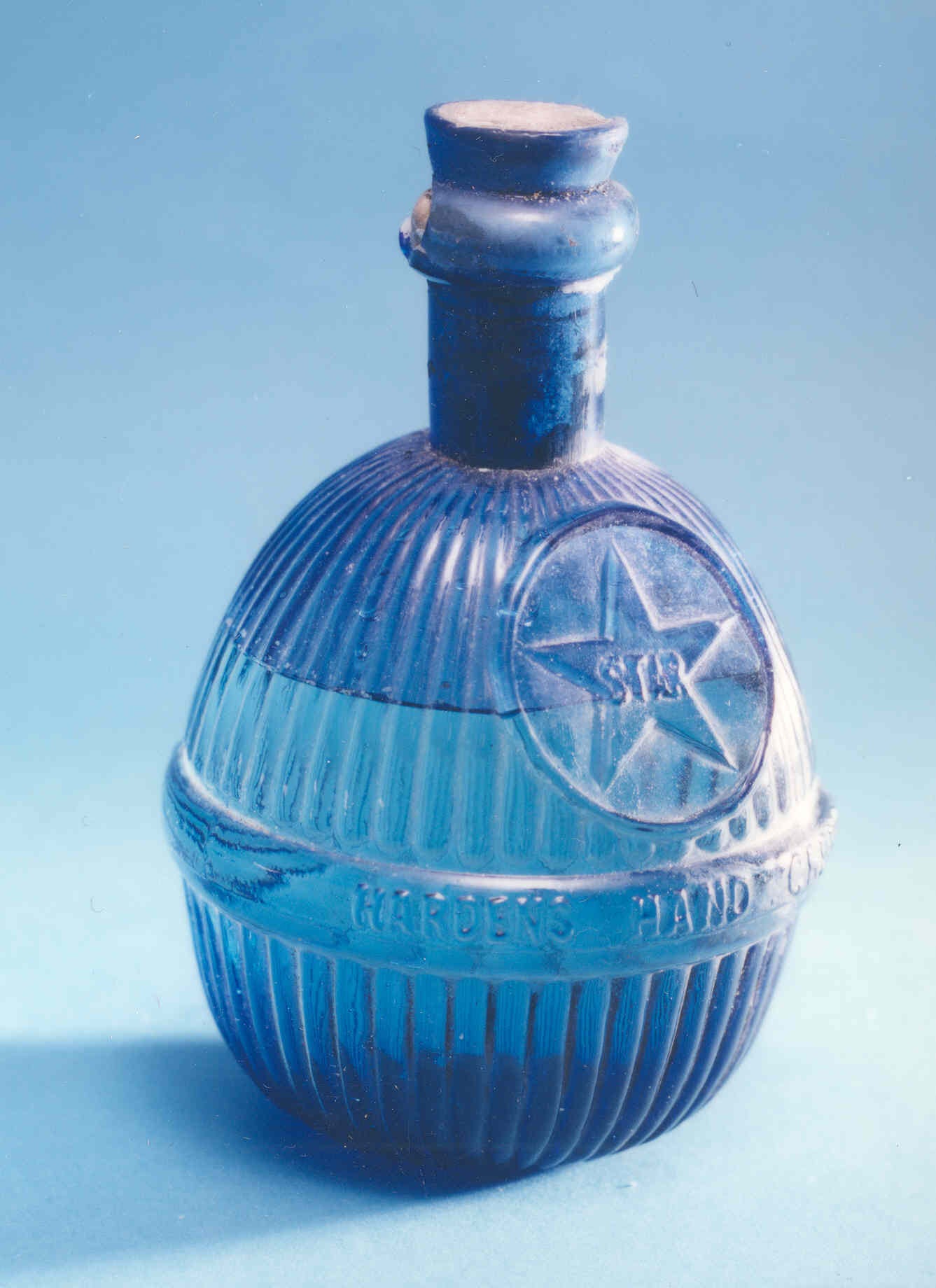 A deep blue coloured glass bottle, almost spherical at the bottom with a short neck on the top. It is filled with liquid and the neck is permanently plugged for safety. The bottle is of ribbed design with a circled star on the front.