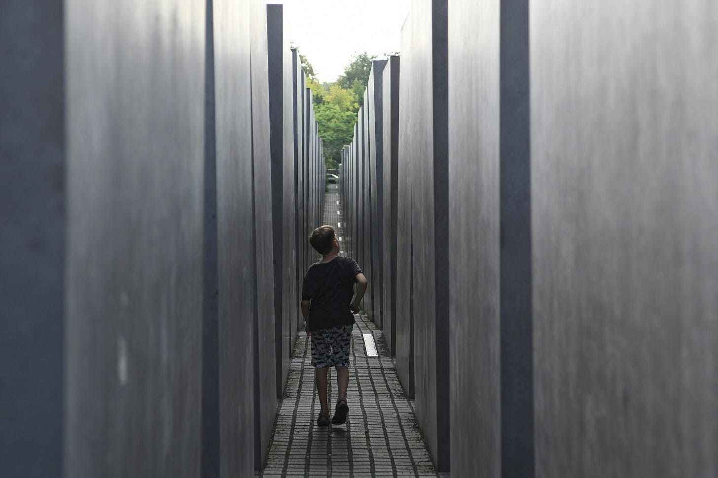 A young boy walks alone down a narrow path flanked by tall, gray concrete blocks. In the distance, a fence and green trees symbolize a distant safe haven.