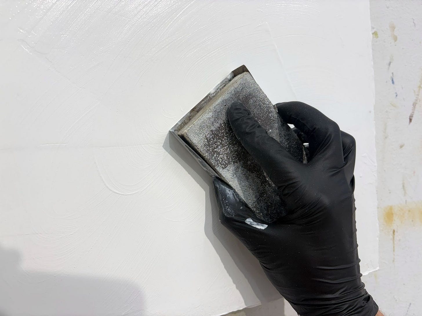 a hand in a black nitrile glove holds a sanding block and appears to be sanding a smooth white surface.