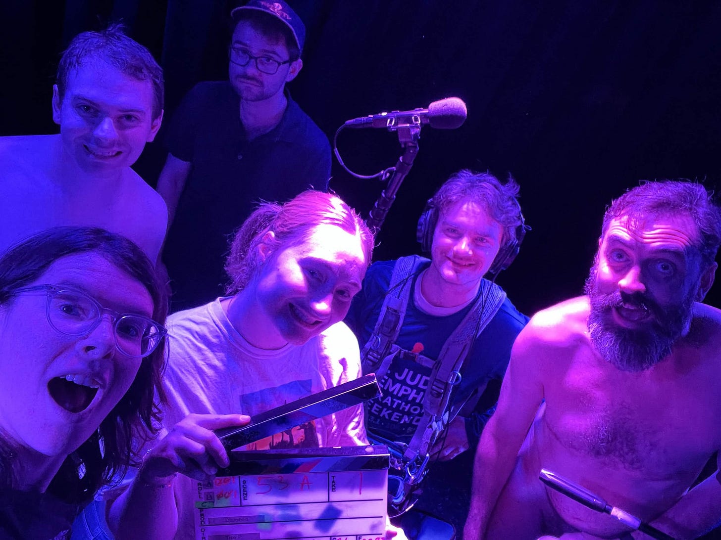 A small group of cast and crew gather closely for a photo under purple stage lighting, smiling and reacting playfully. One person holds a clapperboard in front, while another wears sound equipment with a boom microphone visible overhead. The dark background suggests a stage or performance space during a night shoot.