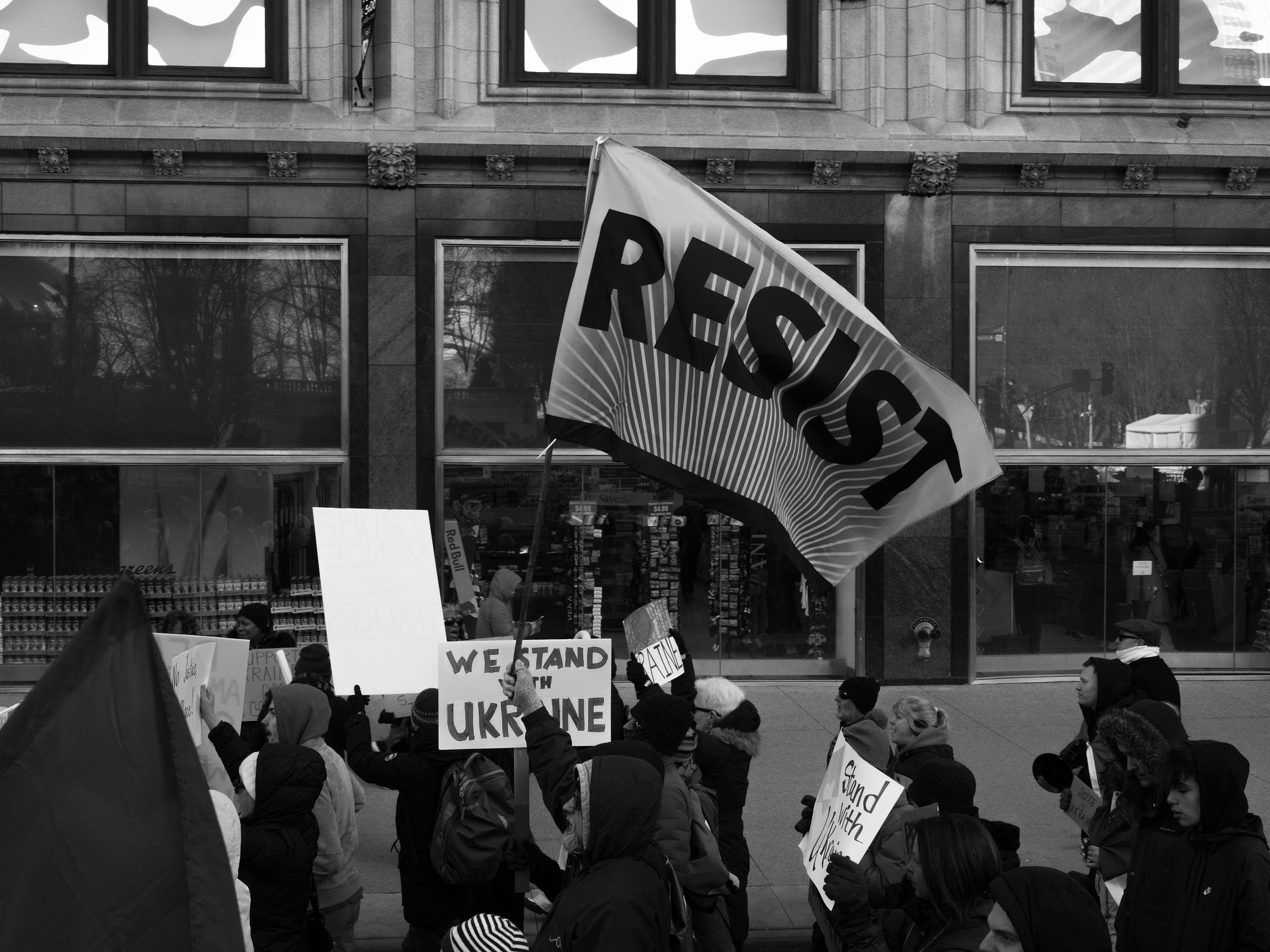 A black and white photograph captures a protest with a large flag reading "RESIST" waving above the crowd. Demonstrators hold various signs, including "We Stand with Ukraine" and "Stand with Our Allies." The protest takes place in front of a city storefront with reflective windows, adding depth to the scene. The flag’s bold lettering and dynamic movement emphasize the spirit of resistance. A black and white photograph captures a protest with a large flag reading "RESIST" waving above the crowd. Demonstrators hold various signs, including "We Stand with Ukraine" and "Stand with Our Allies." The protest takes place in front of a city storefront with reflective windows, adding depth to the scene. The flag’s bold lettering and dynamic movement emphasize the spirit of resistance.