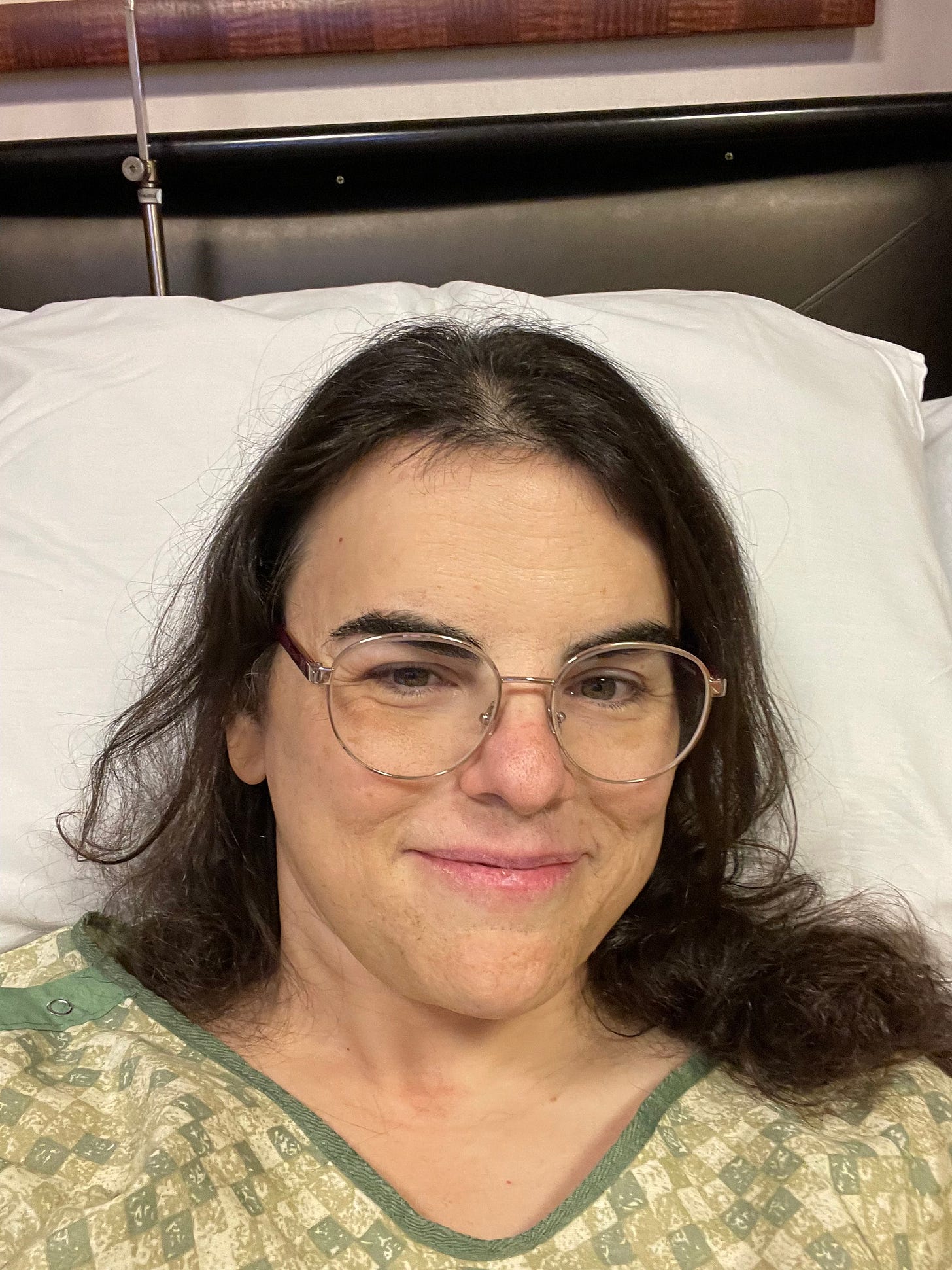 A woman with long black hair and glasses, in a hospital bed, wearing a green hospital gown.