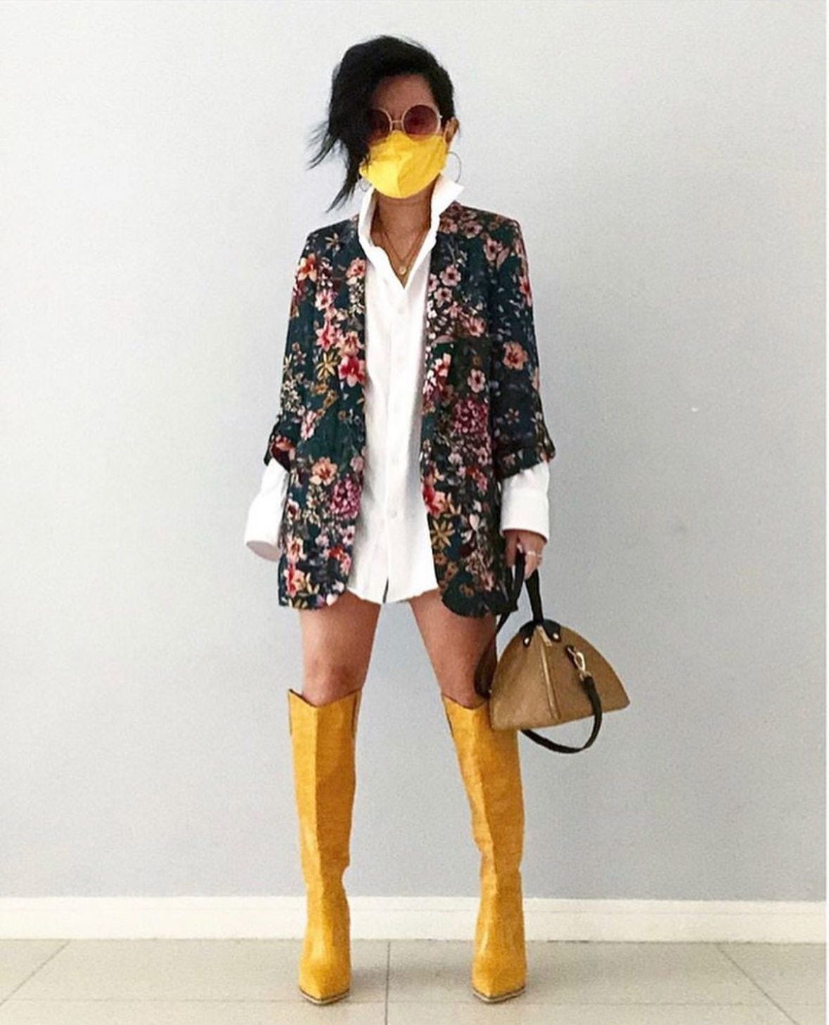 Cancer survivor Sarah in floral blazer, white shirt, yellow boots, and mask holds a tan bag. Cancer survivor Sarah in floral blazer, white shirt, yellow boots, and mask holds a tan bag.