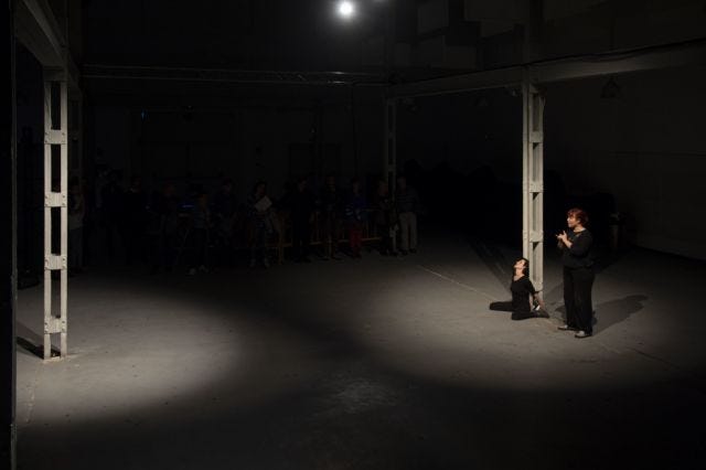 Foto de la performance En cuartas o mitades. Miriam Navarro Prieto de rodillas en el suelo, la espalda apoyada en una columna de metal del antiguo matadero de Valladolid. A su derecha: la intérprete de Lenguaje de signos Inés Alonso. Foto de la performance En cuartas o mitades. Miriam Navarro Prieto de rodillas en el suelo, la espalda apoyada en una columna de metal del antiguo matadero de Valladolid. A su derecha: la intérprete de Lenguaje de signos Inés Alonso.