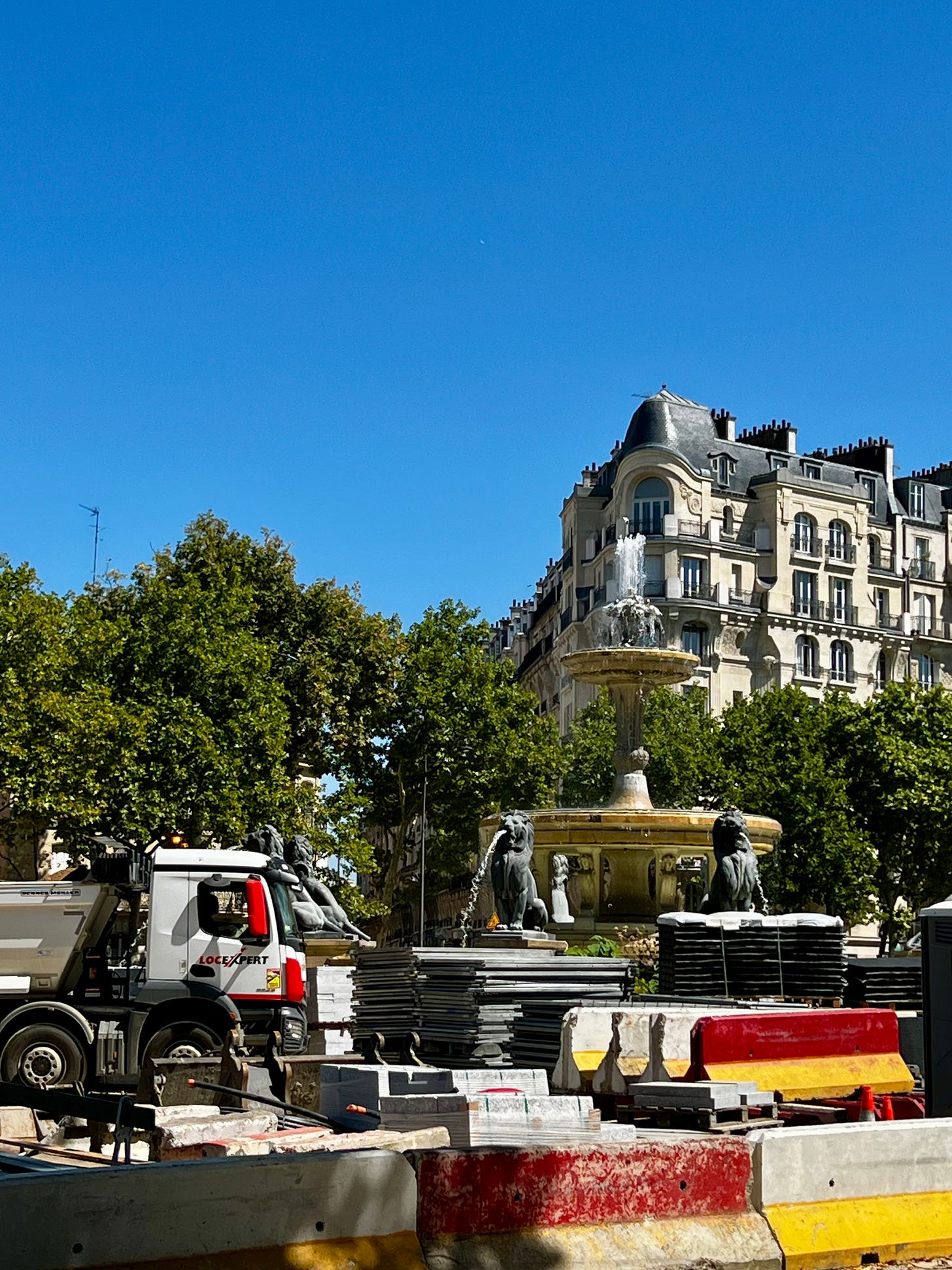 Ornate Parisian fountain surrounded by construction equipment and barriers under bright blue sky.