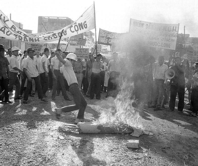 A banner-waving crowd is gathered around a man who is kicking an effigy burning on the ground. A banner-waving crowd is gathered around a man who is kicking an effigy burning on the ground.