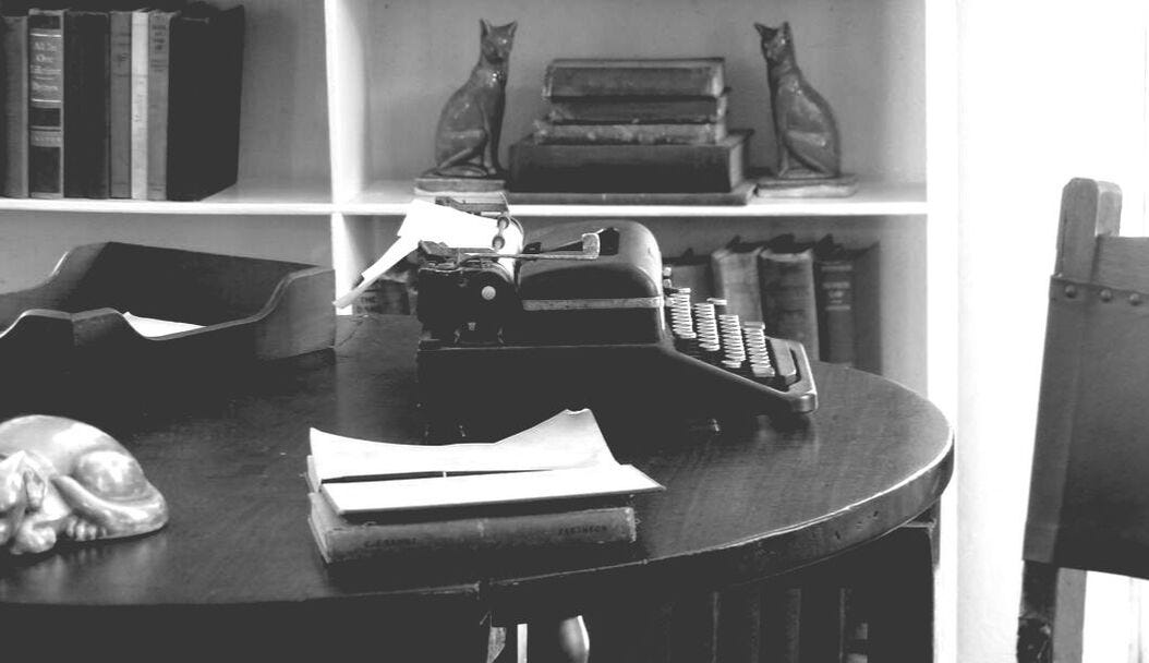 Black‑and‑white photo of a vintage writing desk with an old typewriter, a sheet of paper inserted, a small stack of papers, and a reclining cat figurine. Behind the desk is a bookshelf with neatly arranged books and two matching cat sculptures.