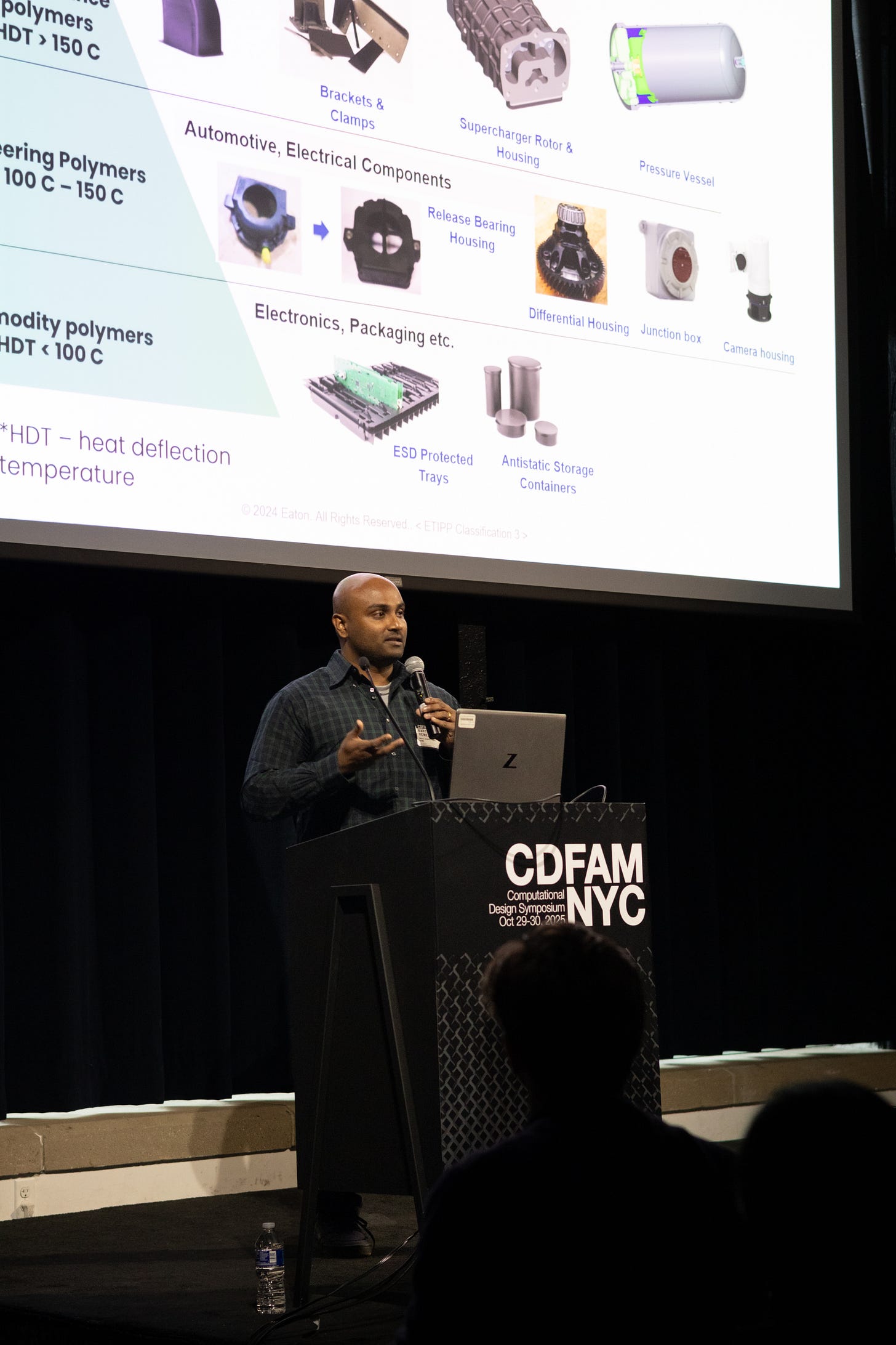 A speaker presenting at a conference event, gesturing towards a projection screen that displays information about various polymers and their applications in industries like automotive and electronics. The backdrop includes a banner for CDFAM NYC. A speaker presenting at a conference event, gesturing towards a projection screen that displays information about various polymers and their applications in industries like automotive and electronics. The backdrop includes a banner for CDFAM NYC.