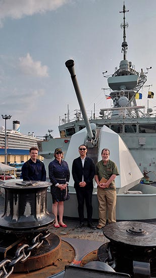 Assistant Deputy Minister (Public Affairs) Geneviève Binet and delegation members stand on the deck of HMCS Charlottetown in front of a naval gun during a visit.