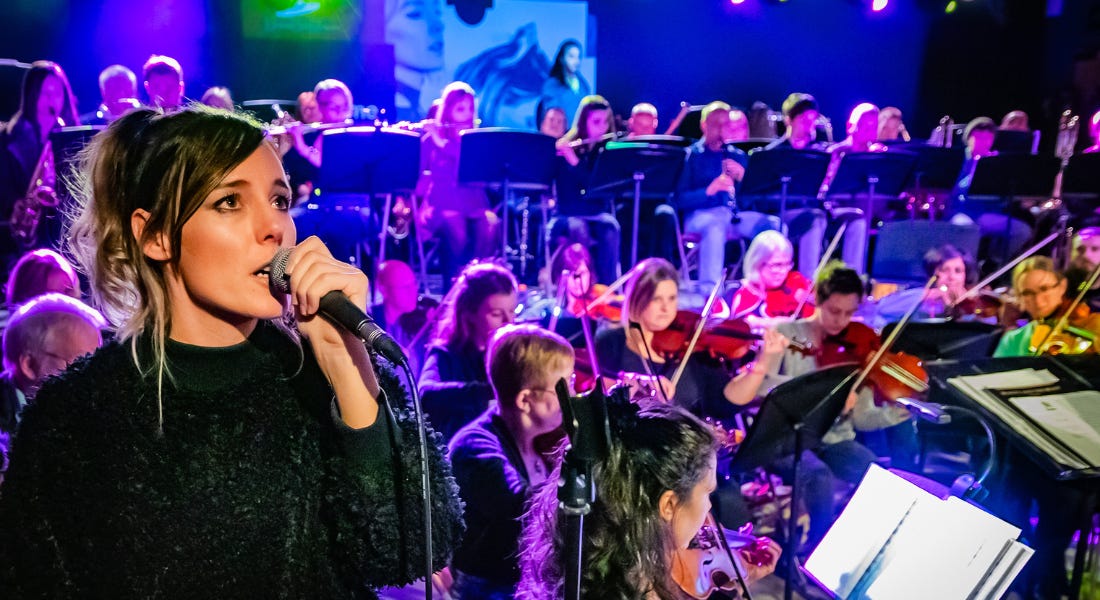 A woman in a black jumper singing into a microphone with an orchestra behind her A woman in a black jumper singing into a microphone with an orchestra behind her