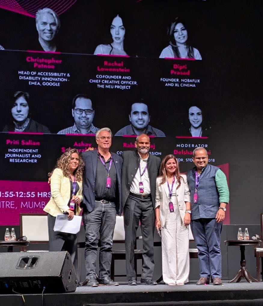 Four panellists stand together on stage with the moderator. From left to right: Delshad, an Indian woman with curly hair wearing a pale yellow blazer over a black outfit, holding papers and smiling; Christopher, a tall white man with short grey hair wearing glasses, a dark blazer over a black shirt, and jeans; Brij, a tall Indian man with a receding hairline and grey beard wearing a dark suit and a white shirt; Dipti, a woman with long dark hair wearing a grey pantsuit and heels, smiling; and Ashay, a man with a receding hairline wearing glasses, a blue vest layering a green linen shirt, and jeans. All five wear purple conference lanyards. The backdrop displays headshots and titles of other speakers.