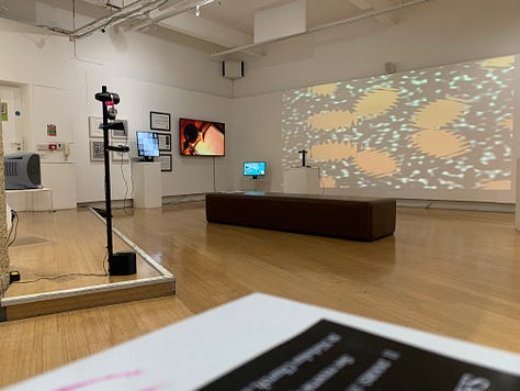 A variety of new media art works using screens, projections, including a large yellow screen and an anaologe cassete with the caption "an actual AI artist" 