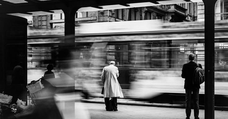This may contain: two people waiting for the train to arrive at the station in black and white photo