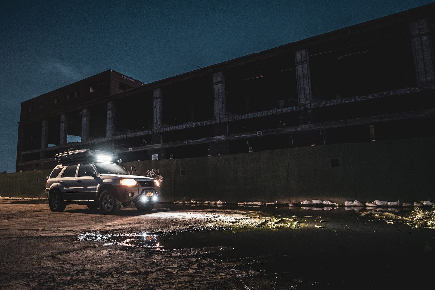 The front of a dark-colored Ford Escape parked on a street in Red Hook, Brooklyn at night. The car's headlights are on, casting a warm glow on the ground in front of it, and the windows are dark.