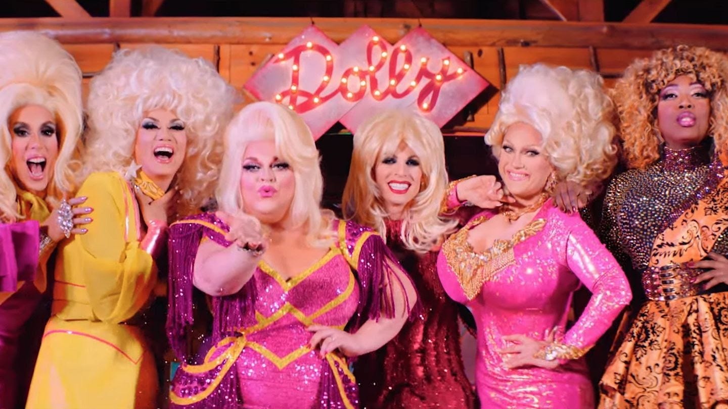Photo of six drag queens dressed in pink and yellow sequin gowns with blonde wigs. They are standing in front of a pink sign that reads "Dolly" in cursive with lights. 
