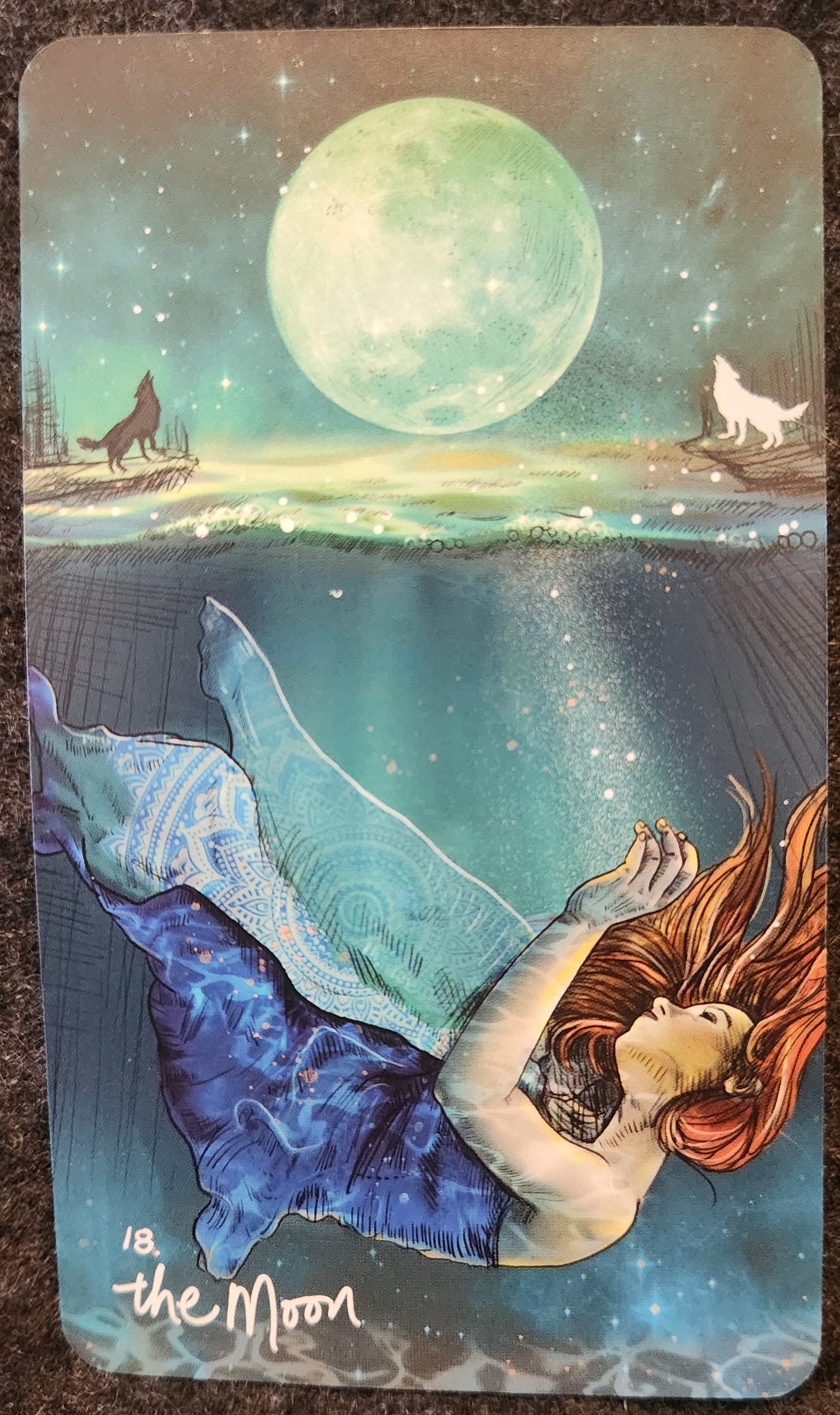 A tarot card labelled 18. The Moon. A woman in a dark blue dress is sinking beneath the water of a moonlit lake. A full moon fills the night sky above. On the left, there is a black dog howling up at the moon. On the right, there is a white wolf howling up at the moon. 