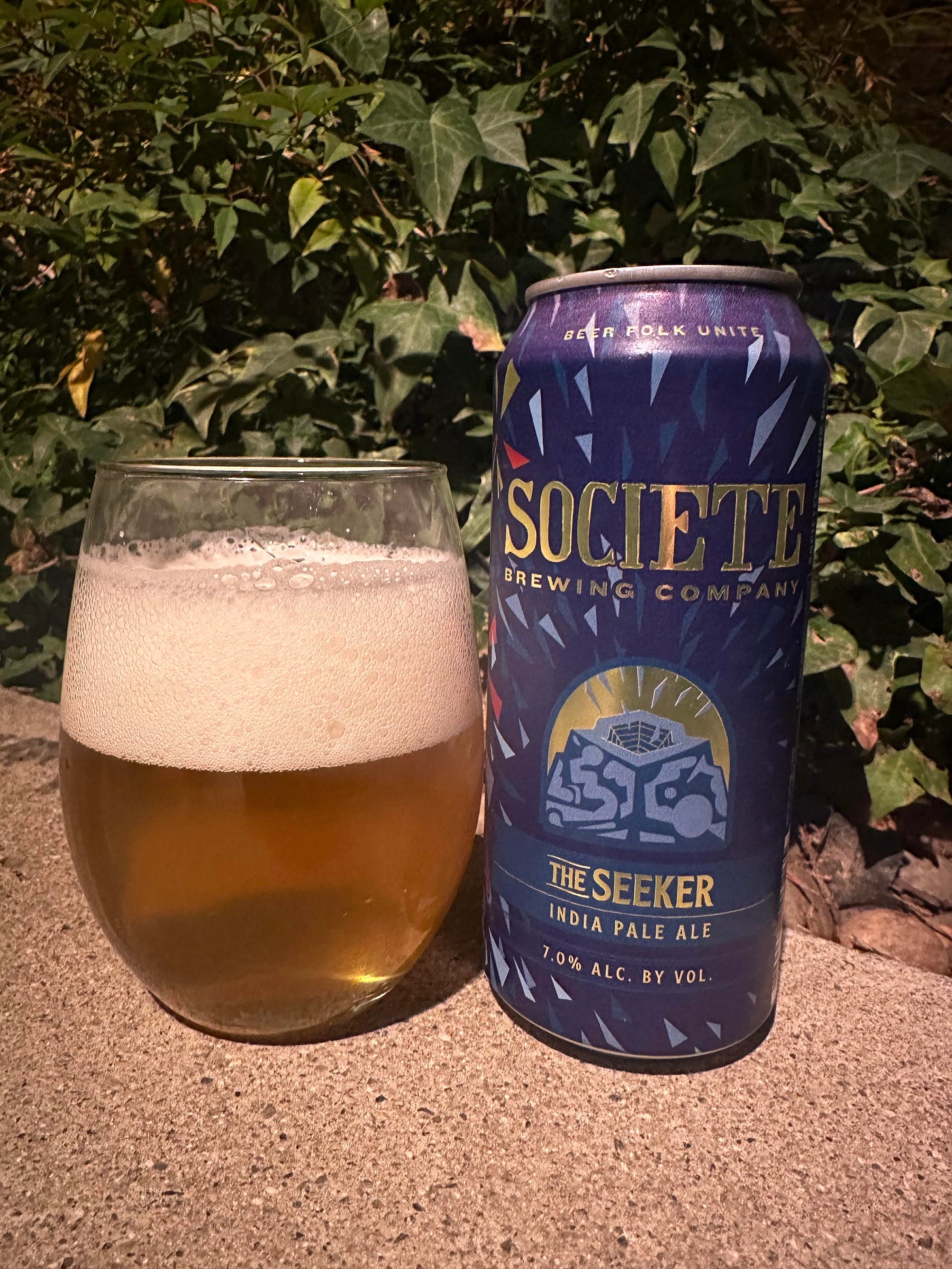 A partially filled beer glass with a foamy head next to a dark blue can of "Seeker IPA" from Societe Brewing