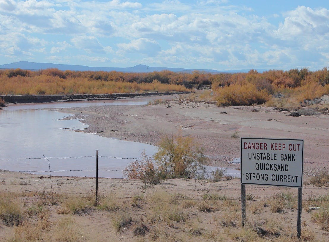 A photo of mountains and blue sky with clouds. In the foreground is a sandy bank and a barbed wire fence. In the lower right corner is a sign that says "Danger keep out unstable bank quicksand strong current."
