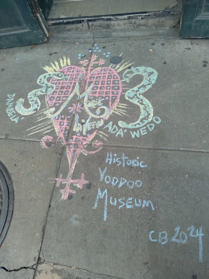 A veve and the words "Historic Voodoo Museum CB 2024" drawn in chalk on a sidewalk. A veve and the words "Historic Voodoo Museum CB 2024" drawn in chalk on a sidewalk.