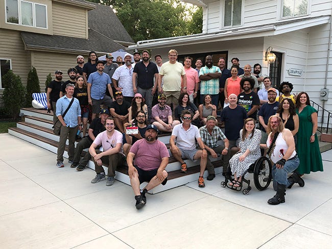 Andy Richter and the cast and crew pose on a patio and set of steps behind a white house. 