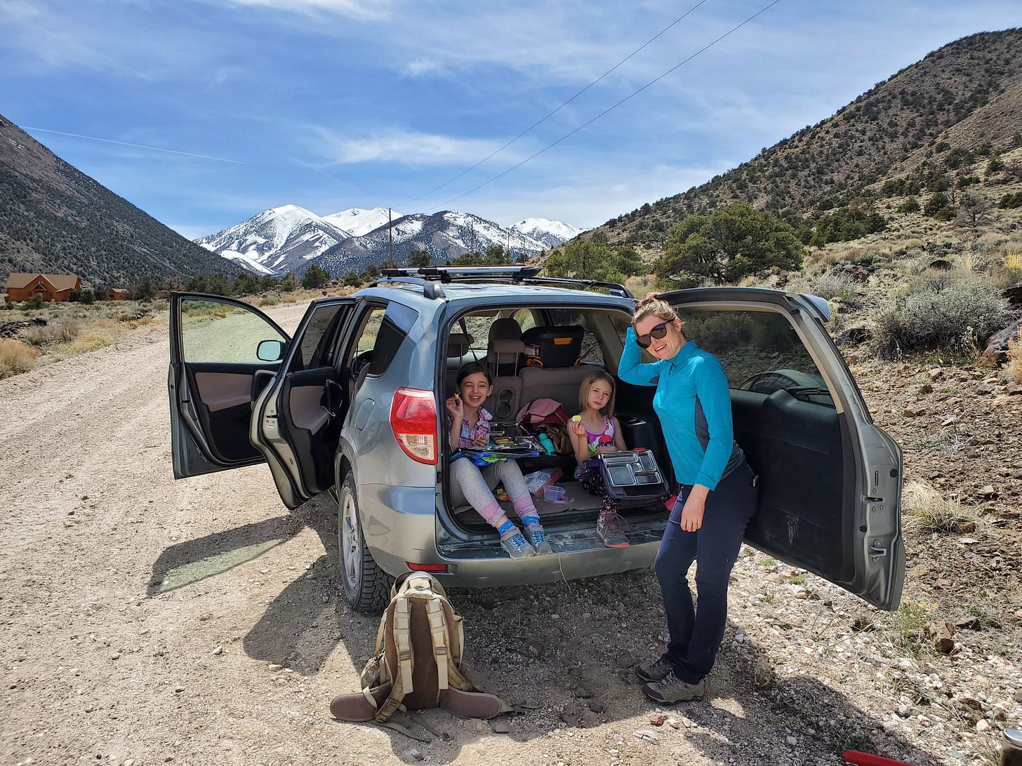 C and L sitting in the back of the car eating lunch. Charlotte standing next to them. Snow capped White Mountains in distance.