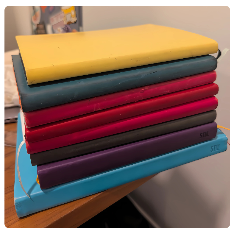 A stack of colorful journals—yellow, teal, red, black, purple, and blue—resting on a wooden desk, showing signs of frequent use.