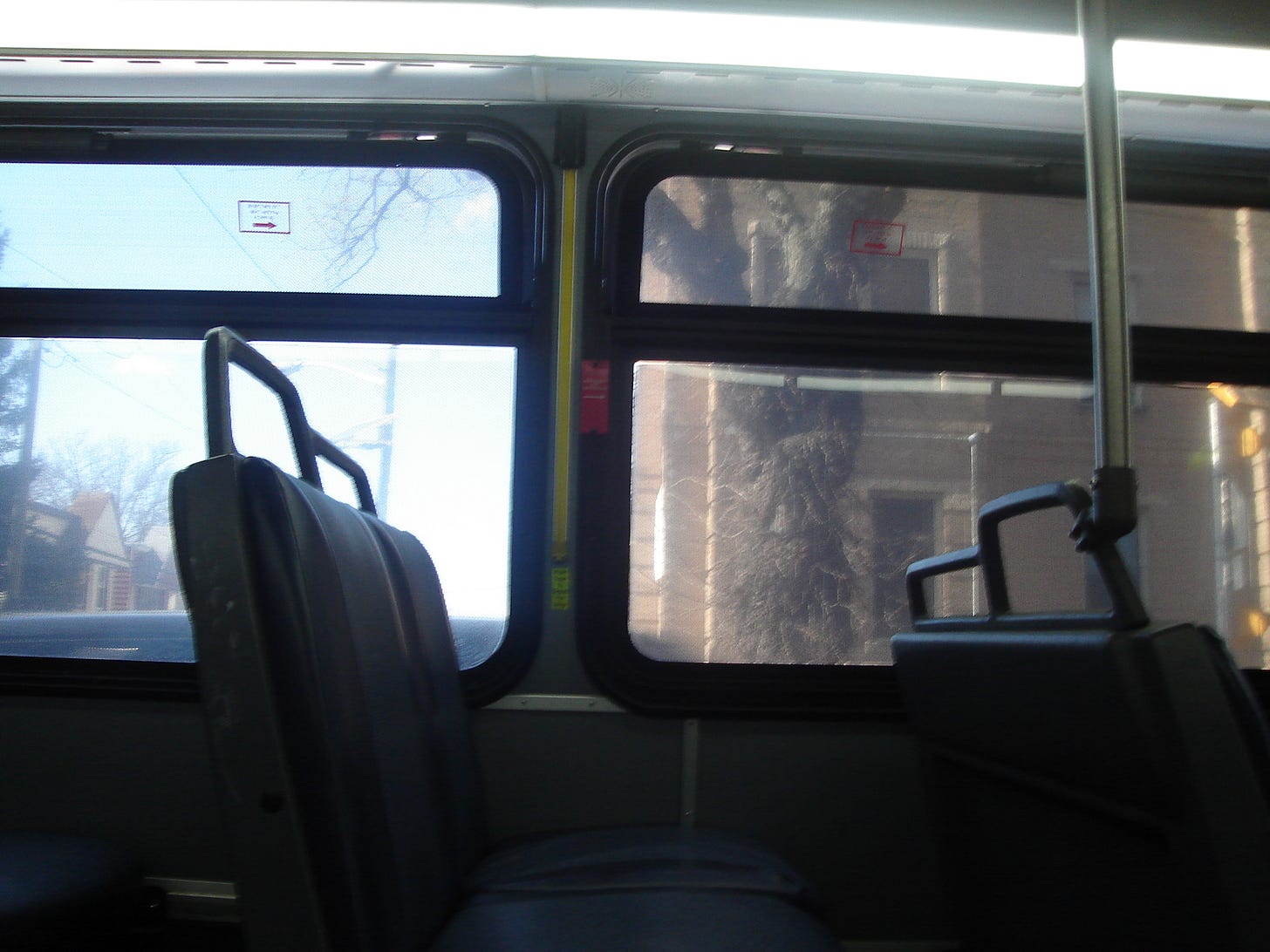 Side view of an empty seat on a bus that highlights that one empty seat in between two partially seen that also look empty. Outside windows, it's a bright day through an urban landscape. A leafless tree is visible in front of a sandy-colored building. 