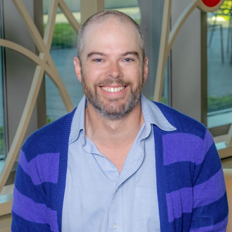 A white man smiling, with shaved hair, wearing a blue dress shirt with purple cardigan.