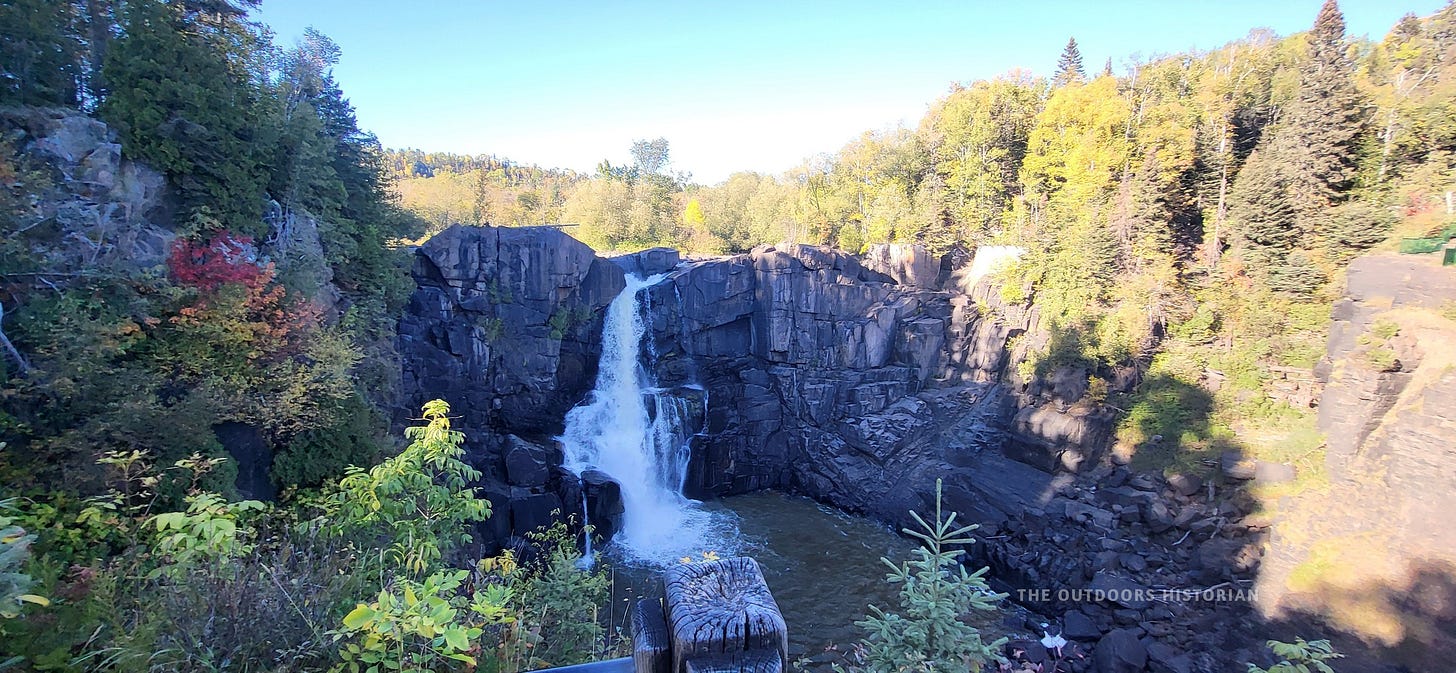 A view of High Falls on the Pigeon River during a sunny late September afternoon. The tall waterfall is partially in deep shadow, with golden autumn light hitting the rushing water and the rocky cliffs.