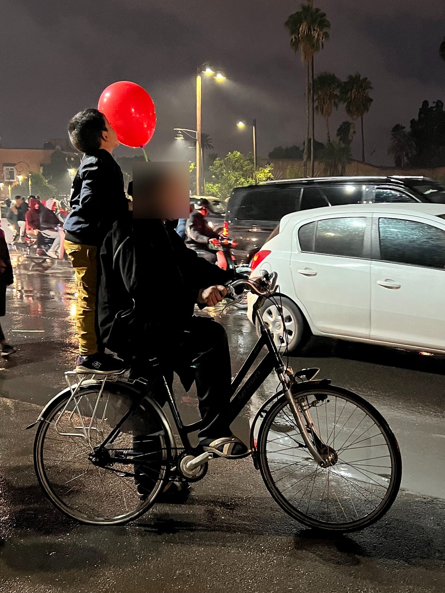 A young boy standing on the back of a bicycle kisses a red Morocco balloon as crowds celebrate a World Cup victory on the rain-soaked streets of Marrakech.