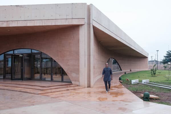 An man walks toward the camera on a wide paved path outside a modern building with curved walls and large arched glass entrance doors.