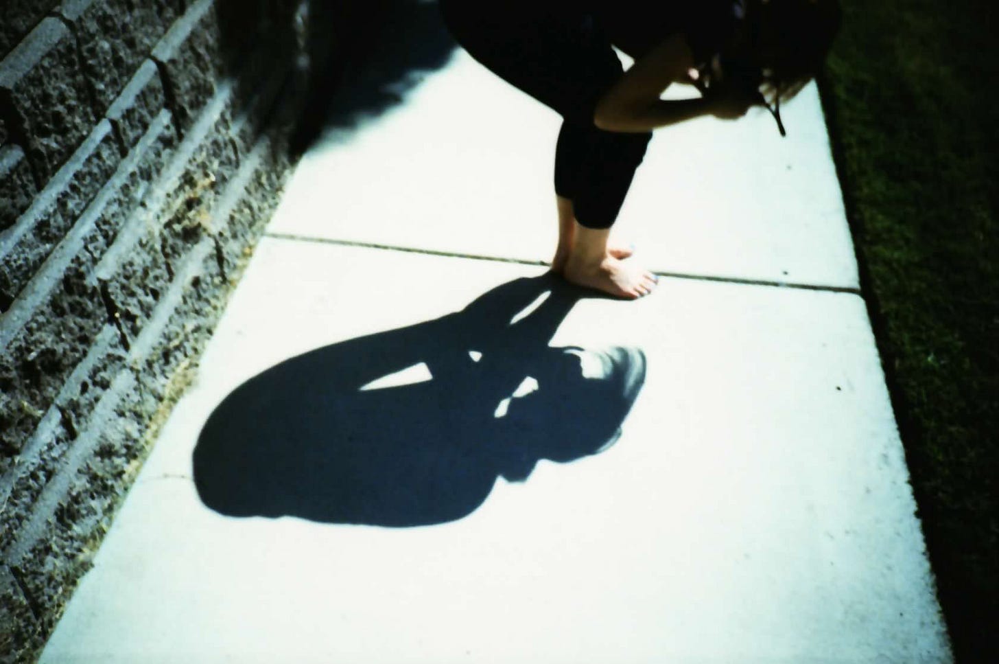 A photograph predominantly of Sarah's shadow, with her body seen near the upper edge of the frame. She is leaning over with a camera and taking a picture of the ground. Standing on the sidewalk, a brick wall can be seen at the left side of the frame.