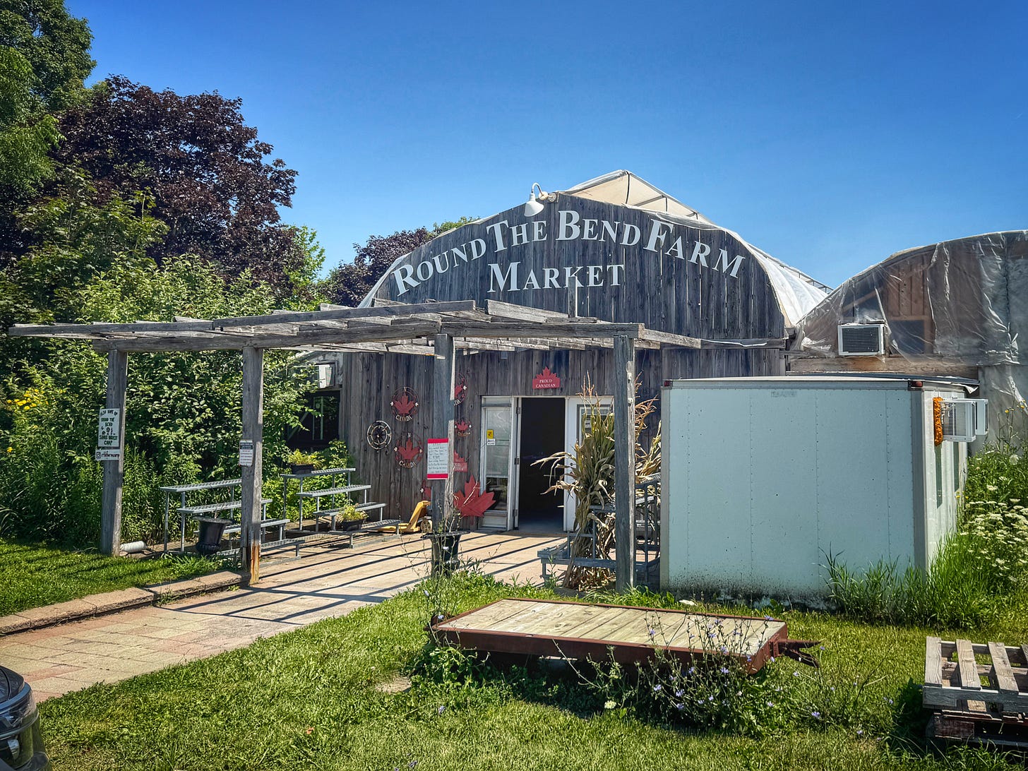 The front of Round the Bend Farm Market. It's a very rustic building with a barn board front and an arched roof that meets in a small point at the top. There's a bare gazebo covering the entrance to the market and empty plant stands beside the front door. The sky is very blue and the grass is well kept. The front of Round the Bend Farm Market. It's a very rustic building with a barn board front and an arched roof that meets in a small point at the top. There's a bare gazebo covering the entrance to the market and empty plant stands beside the front door. The sky is very blue and the grass is well kept.