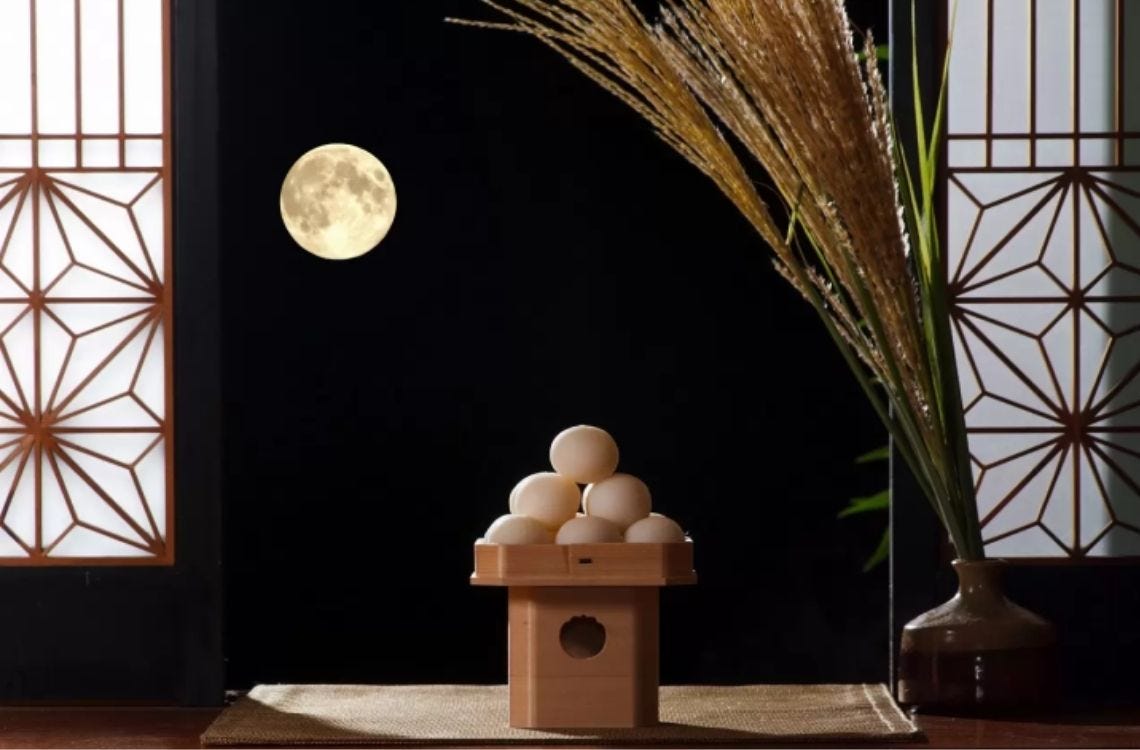 A traditional Japanese tsukimi (moon-viewing) display with pampas grass in a vase, a wooden stand holding stacked rice dumplings, and a full moon visible through sliding shoji doors.