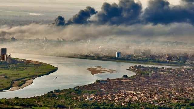 Vast plumes of dark smoke rise above a city situated on a river.