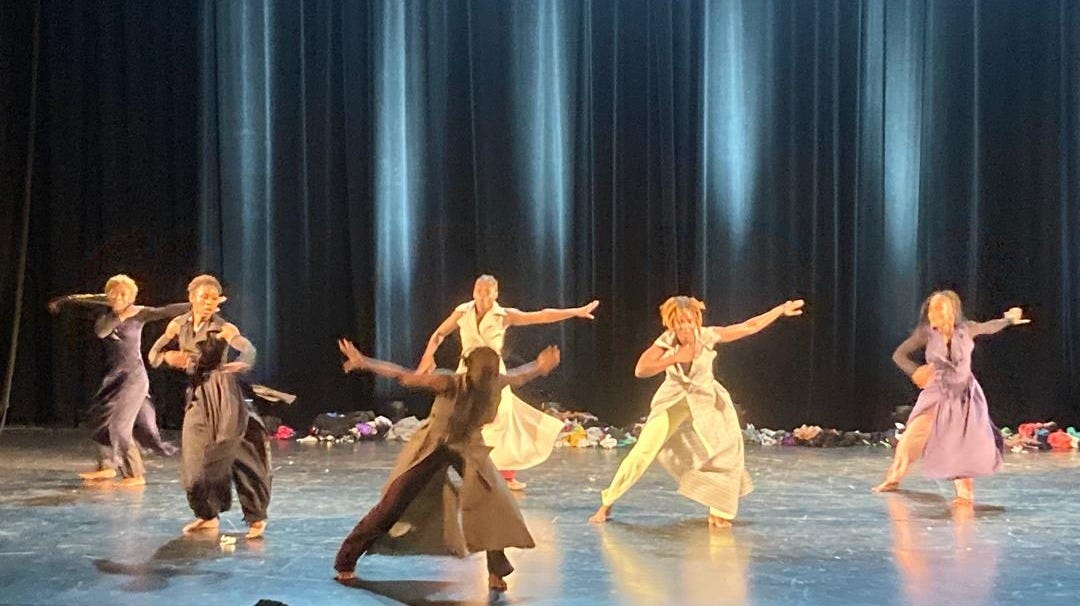 Six Black African dancers wearing colorful dress while dancing on stage. A dark blue curtain shimmers behind them. 