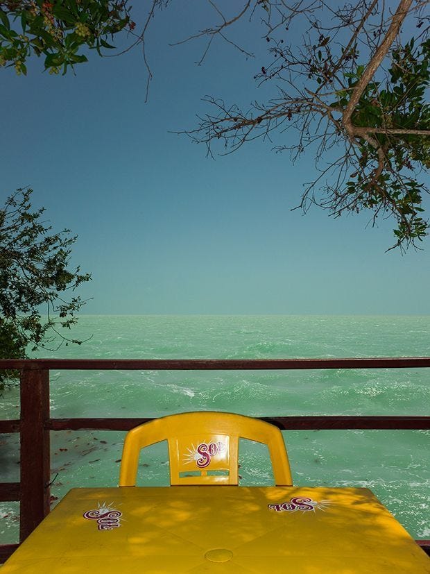 This may contain: a yellow table and chair sitting on a deck overlooking the ocean with trees in the foreground